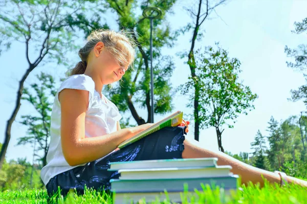 Una chica enseña tareas en libros de texto y se sienta en la hierba verde en el parque . — Foto de Stock