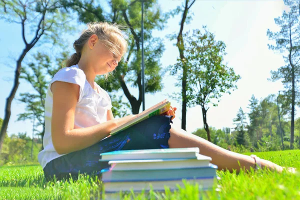 Una chica enseña tareas en libros de texto y se sienta en la hierba verde en el parque . — Foto de Stock