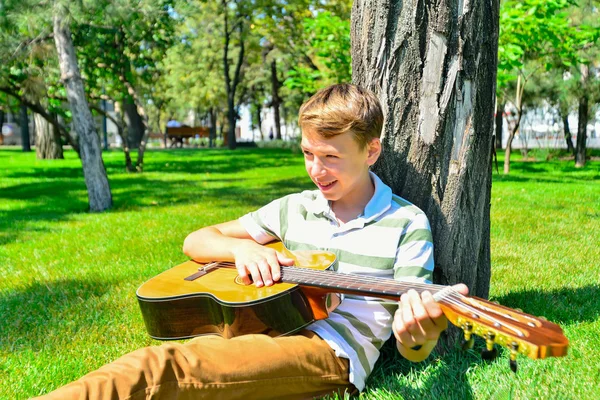 Un chico con una guitarra bajo un árbol, un adolescente toca y canta en el parque . —  Fotos de Stock