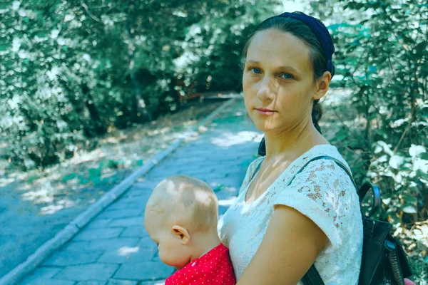 Une maman jeune et heureuse s'assoit avec un bébé dans ses bras sur un banc de parc . — Photo