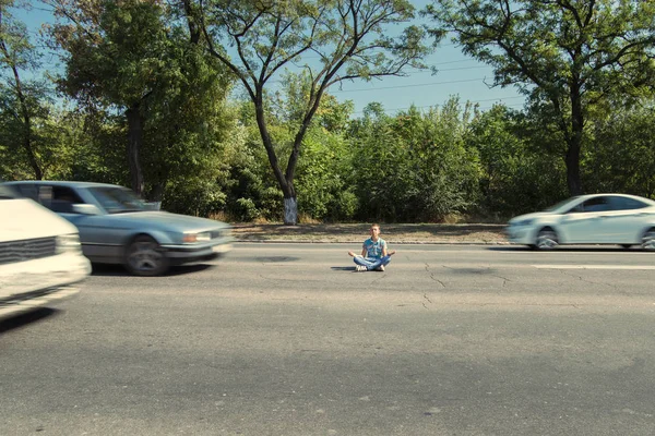 A man sits on an asphalt road and meditates past passing cars — Stock Photo, Image