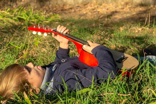 Un niño toca la guitarra tumbado en la hierba verde del parque . —  Fotos de Stock