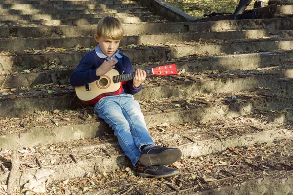 Un niño con chaqueta azul y jeans toca la guitarra en los viejos escalones de un parque abandonado . — Foto de Stock