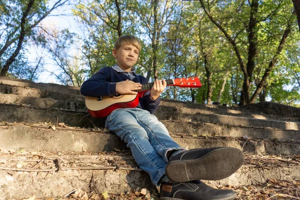 Un niño con chaqueta azul y jeans toca la guitarra en los viejos escalones de un parque abandonado . —  Fotos de Stock