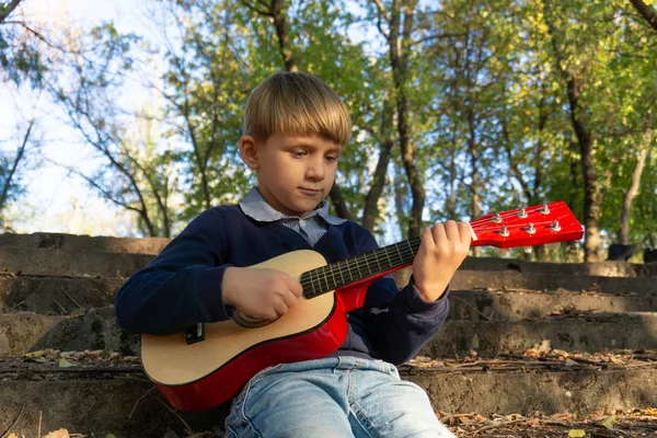 Un niño con chaqueta azul y jeans toca la guitarra en los viejos escalones de un parque abandonado . —  Fotos de Stock