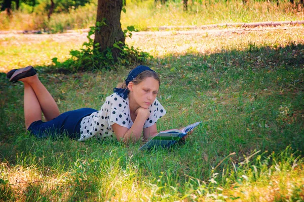 Una chica yace en la hierba debajo de un árbol y lee un libro . — Foto de Stock