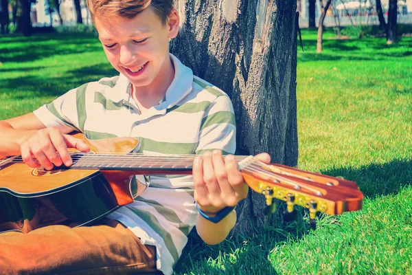 Un chico con una guitarra bajo un árbol, un adolescente toca y canta en —  Fotos de Stock