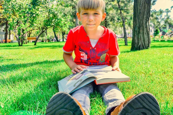 Um menino de roupas vermelhas segura um livro em suas mãos e senta-se no g — Fotografia de Stock