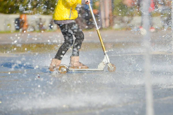 Een meisje in een gele jas paardrijden een scooter in een fontein onder spray van water. — Stockfoto