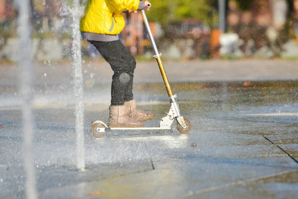 Een meisje in een gele jas paardrijden een scooter in een fontein onder spray van water. — Stockfoto