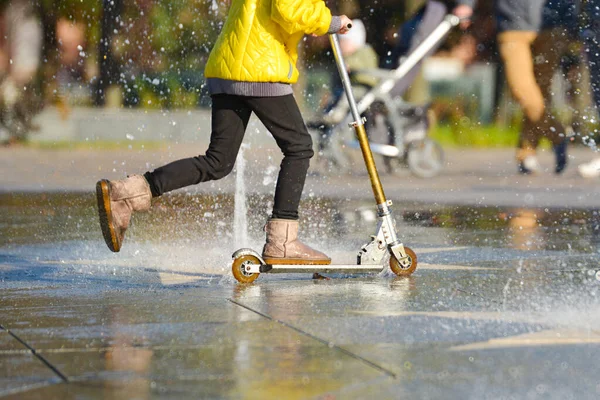 Een meisje in een gele jas paardrijden een scooter in een fontein onder spray van water. — Stockfoto