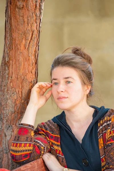 Une fille s'assoit sur un banc dans un parc près d'un arbre et regarde ailleurs . — Photo