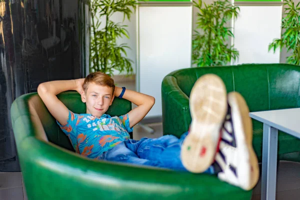 O menino se senta em uma poltrona verde, pernas encimadas e desfruta do resto, olhando para a câmera . — Fotografia de Stock