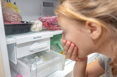 A surprised girl looks at the full ice cube tray of the freezer ice maker. clipart