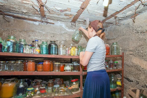 The woman in the cellar is gray-haired, the girl prepares food for the winter, canned food in glass jars on the rack. — Stock Image Woman Cellar Gray Haired Girl Prepares Food Winter Canned Food Stock Picture