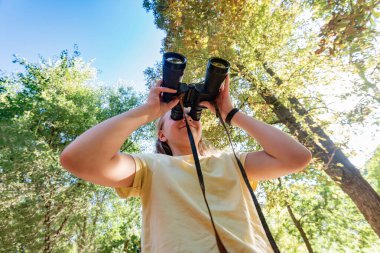 Yürüyüşe çıkmış bir kız, ormanın ortasında yolun ortasında dürbünle bakıyor..