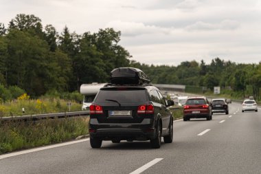 Çatı kutusu olan siyah bir SUV çok şeritli bir otoyolun sağ şeridinde gidiyor. Arka planda yeşil ağaçların sıralandığı diğer araçlar da arkadan takip ediyor.