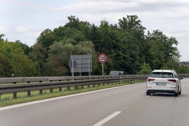 Otoyol boyunca hız limiti levhası ile işaretlenmiş beyaz bir araba saatte 120 kilometre yol alıyor. Bereketli yeşillik parlak bir gökyüzünün altındaki yolu çiziyor..
