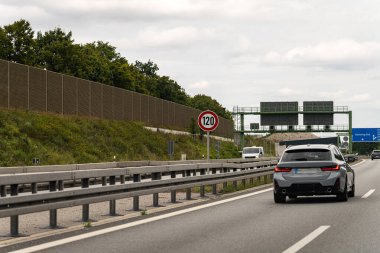 Yeşillik ve gürültü bariyerleriyle dolu bir otoyol boyunca parlak gri bir araba sürüyor. Görünür hız sınırı işareti, Almanya 'da parçalı bulutlu bir günde saatte 120 km hıza işaret ediyor..