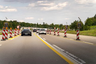 Arabalar, yoğun bir öğleden sonra bulutlu bir gökyüzünün altında uyarı işaretleri ve trafik bariyerleriyle çevrili bir otoyoldaki inşaat alanından geçerler..