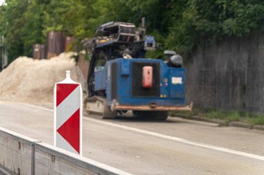 Kırmızı ve beyaz işaretli bir trafik işareti yol çalışma alanını gösteriyor. Arka planda, mavi bir inşaat aracı, bu kentsel alanda devam eden projeler için malzeme yığınının yanında çalışıyor..