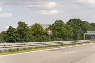 Bir otoyolun kenarında hız limiti tabelası saatte 80 km 'yi gösteriyor. Yol boyunca uzanan yemyeşil ağaçlar, arka planda parçalı bulutlu bir gökyüzüne karşı bir bina görülebilir..