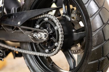 Detaylı bir görüş, motorsikletin zincirini, arka dişlisini ve iyi aydınlatılmış bir atölyede fren diskini yakalar. Bu, genellikle motosiklet bakımında bulunan karmaşık mekanikleri vurgular.