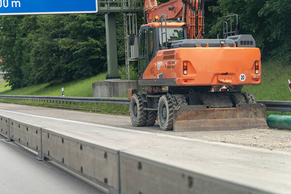 Тяжелая строительная машина припаркована рядом с шоссе, работая на поверхности дороги, в то время как движение в фоновом режиме в течение дня. Зеленые деревья окружают местность.