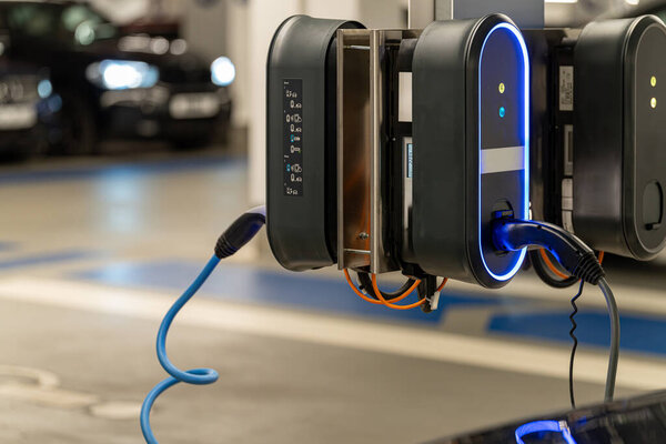 Два электрических зарядных устройства устанавливаются на стене в крытом гараже. Они активно заряжают транспортное средство, в то время как рядом припаркован другой автомобиль. Окружающая среда чистая и современная.