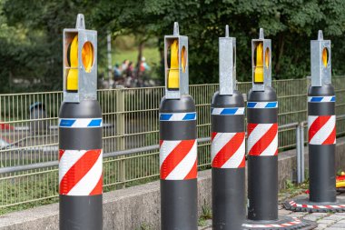 Uyarı ışıklarıyla birlikte birkaç trafik güvenlik direği canlı kentsel bir bölgede bir patika boyunca yerleştirilmiştir. Güneşli bir günde arka planda bisikletçiler ve yayalar görülebilir.