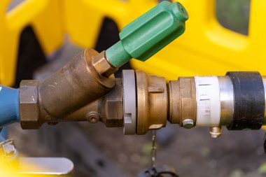 Makinelere bağlı metalik bir kapakçığa yakından bakarak yeşil kabzayı vurguluyor. Çevrede devam eden çalışmalara işaret eden sarı inşaat ekipmanları bulunuyor..