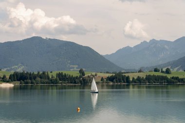 Bir yelkenli bulutlu bir gökyüzünün altında sakin bir göl boyunca usulca hareket eder. Verimli yeşil tepeler ve görkemli dağlar yaz boyunca güzel bir zemin oluşturur..