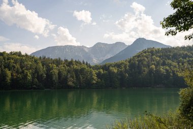 Sakin bir göl, yüksek dağları ve canlı yeşilliği yansıtır. Suyun üzerinde süzülen küçük bir tekne. Güneş ışığı huzurlu ortamı aydınlatır..