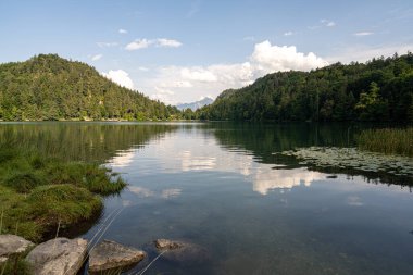 Sakin bir göl etrafındaki dağları ve ağaçları yansıtır. Yumuşak bulutlar mavi gökyüzünde süzülür, sakin bir yaz atmosferi yaratır. Işıldayan su doğal güzelliği artırır..