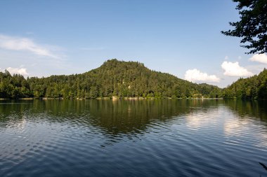 Yoğun ormanlarla çevrili sakin bir göl ve belirgin bir tepe manzarayı yakalar. Yumuşak bulutlar mavi gökyüzünde süzülür ve akşam erken saatlerde sakin bir atmosfer yaratır..