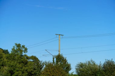 Güç hatları canlı mavi gökyüzüne karşı yükseliyor, yeşil ağaçlarla çevrili, huzurlu bir kırsal alanda. Parlak güneş ışığı sakin atmosferi vurgular.