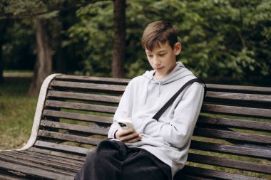Teenage boy sitting on a park bench outdoors, looking at his smartphone. Relaxed urban lifestyle, youth activity, technology use, and casual leisure concept.