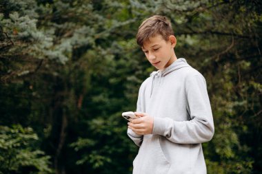Teenage boy outdoors in casual clothing in a forest, looking at his smartphone. Concept of gadget addiction, digital lifestyle, youth, and modern mobile dependency during a walk. Copy space.
