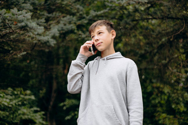 Portrait of a serious teenage boy outdoors in a park, talking on his smartphone. Concept of remote communication, staying connected, youth, and modern urban lifestyle.