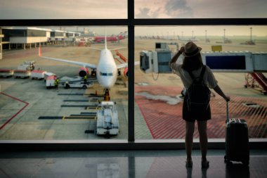 Young asian woman with baggage looking through the window of an airport departure lounge