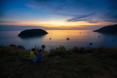 Romantic scene of middle aged love couple sitting on grass in view point look sunset at Promthep Cape Phuket, Thailand.