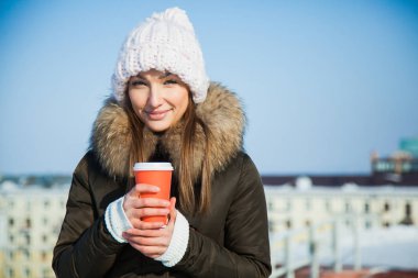 Güneşli soğuk soğuk kış gününde gitmek sevimli genç bayan ile kahve. Büyük rahat Yün şapkalı kadın sıcak içecek donmuş bilek ısınma tutar.
