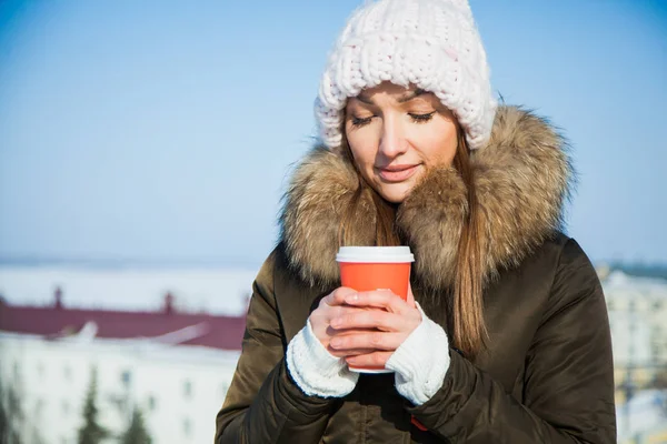 Cute young lady with coffe to go on a sunny frosty cold winter day. Woman in big cozy woolen hat holds hot drink warming frozen wrists.