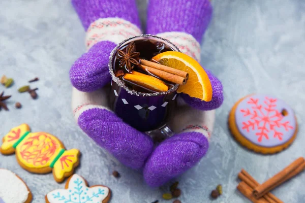 Mulled wine in glass mug with spices, female hands in knitted mittens. Christmas hot drink with decorated new year ginger bread on gray stone table. Top view.