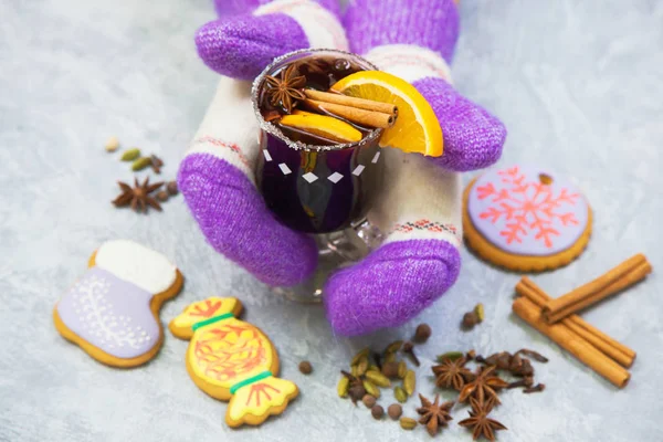 Mulled wine in glass mug with spices, female hands in knitted mittens. Christmas hot drink with decorated new year ginger bread on gray stone table. Top view.