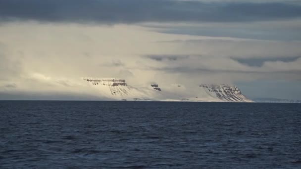 Snötäckta klippor i dimman, upplysta av solen vid horisonten. I förgrunden är det blå havet — Stockvideo