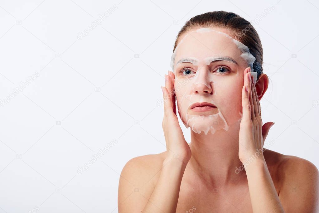 Beautiful young caucasian woman applying paper sheet mask on her face isolated on white background. Beauty skin care concept. Cosmetology.