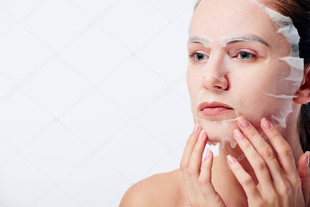 Beautiful young caucasian woman applying paper sheet mask on her face isolated on white background. Beauty skin care concept. Cosmetology.
