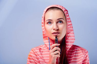 Hood ve kulaklık üzerinde mavi arka plan izole vape sigara ile parlak ceketli genç bir kızın portresi. Buharı kavramı. Modern eğilim vaping e-sigara.