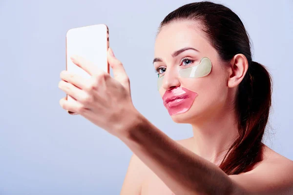 Beautiful woman with natural make-up, blue hyaluronic acid hydrogel patches and pink lips mask taking a selfie with her smartphone. Korean cosmetics. Cosmetology and skin care. Isolated on grey